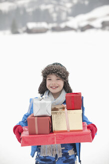 Porträt eines lächelnden Jungen mit einem Stapel Weihnachtsgeschenke im Schnee - CAIF12421