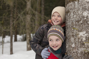 Happy boys leaning against tree trunk in snowy woods - CAIF12424
