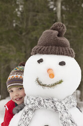 Porträt eines lächelnden Jungen hinter einem Schneemann mit Strumpfmütze - CAIF12428