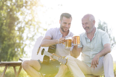 Vater und erwachsener Sohn stoßen mit Bierkrügen an und spielen Gitarre im Freien - CAIF12680