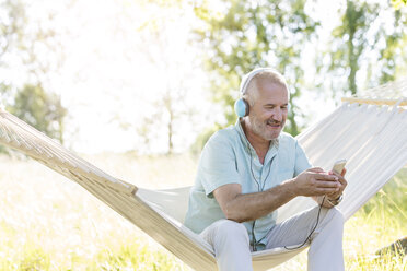 Älterer Mann mit Kopfhörern, der mit einem mp3-Player in einer Sommerhängematte Musik hört - CAIF12681