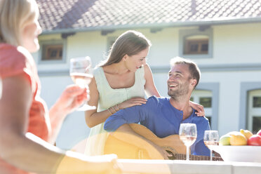 Lächelndes junges Paar spielt Gitarre und trinkt Wein auf einer sonnigen Veranda - CAIF12701