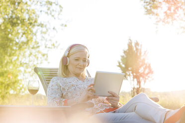 Ältere Frau mit Kopfhörern und Wein, die auf einem Liegestuhl im sonnigen Hinterhof ein digitales Tablet benutzt - CAIF12721