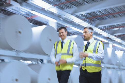 Businessmen in reflective clothing walking along large paper spools in printing plant - CAIF12969