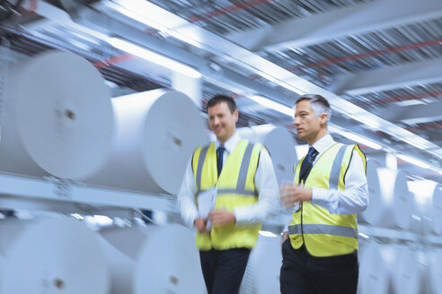 Geschäftsleute in reflektierender Kleidung gehen entlang großer Papierrollen in einer Druckerei - CAIF12969