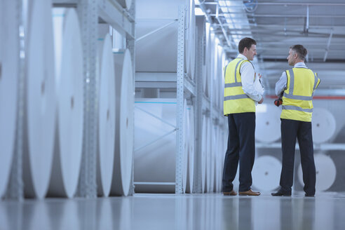 Businessmen in reflective clothing talking near large paper spools in printing plant - CAIF12983