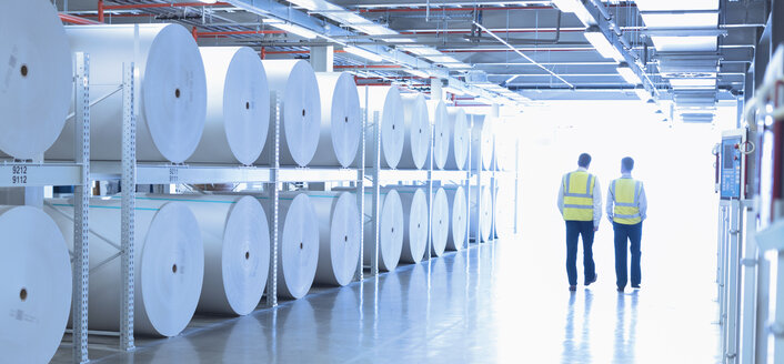 Arbeiter in reflektierender Kleidung, die in einer Druckerei an großen Papierspulen entlanggehen - CAIF12989