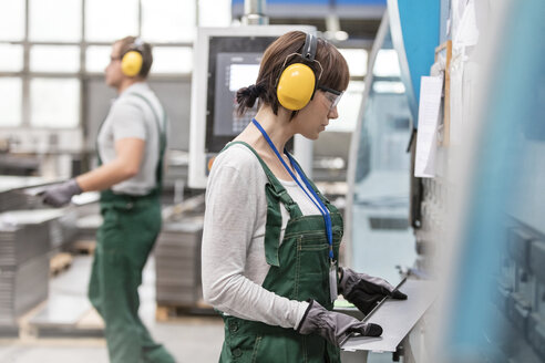 Weiblicher Arbeiter mit Gehörschutz hält Metallteil in Fabrik - CAIF13108