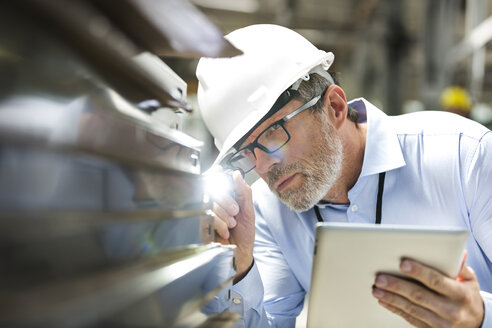 Focused engineer with digital tablet and flashlight examining part in factory - CAIF13158