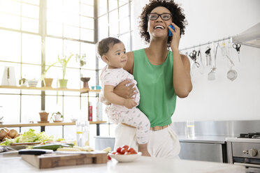 Mutter hält ihre kleine Tochter, während sie in der Küche ein Mobiltelefon benutzt - CAIF13296