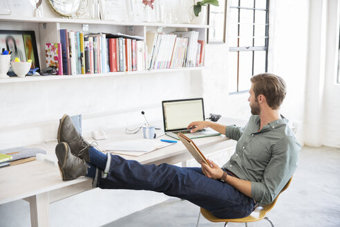 Porträt eines jungen Mannes, der mit den Beinen auf dem Schreibtisch sitzt und mit einem Laptop arbeitet - CAIF13304