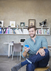 Young blonde man sitting with mobile phone in studio - CAIF13306