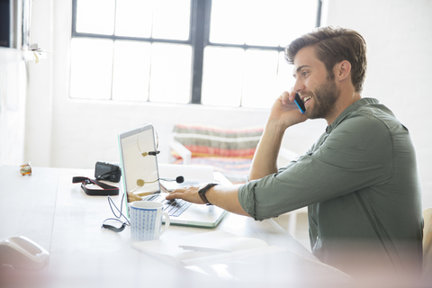 Porträt eines jungen Mannes am Schreibtisch sitzend mit Mobiltelefon und Laptop, lizenzfreies Stockfoto