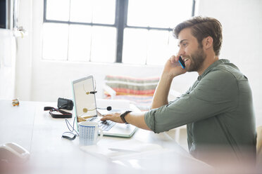 Porträt eines jungen Mannes am Schreibtisch sitzend mit Mobiltelefon und Laptop - CAIF13310