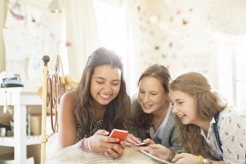 Drei Mädchen im Teenageralter benutzen gemeinsam ein Smartphone, während sie auf dem Bett im Schlafzimmer liegen, lizenzfreies Stockfoto