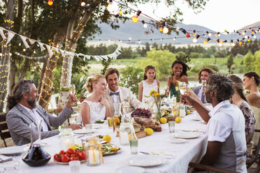 Junges Paar und ihre Gäste sitzen am Tisch während der Hochzeitsfeier im Garten - CAIF13487