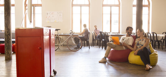 Casual business people working at laptop on bean bag chairs in open office - CAIF13615