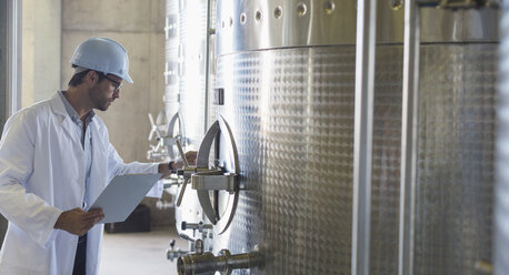 Vintner in lab coat and hard hat examining stainless steel vat in cellar - CAIF13649