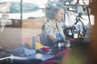 Lächelnde Frau mit Kopfhörern am Laptop in einem Cafe - CAIF13711