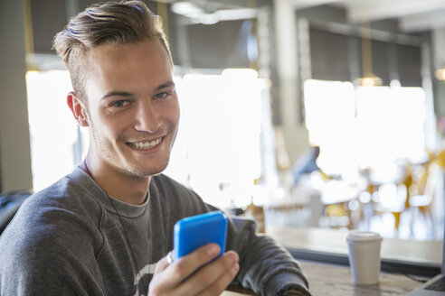 Porträt eines lächelnden jungen Mannes, der in einem Café eine SMS mit seinem Mobiltelefon schreibt - CAIF13724