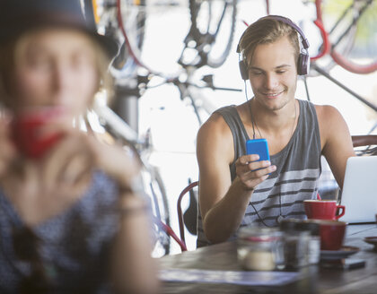 Junger Mann mit Kopfhörern und Laptop, der mit seinem Handy in einem Café SMS schreibt - CAIF13725