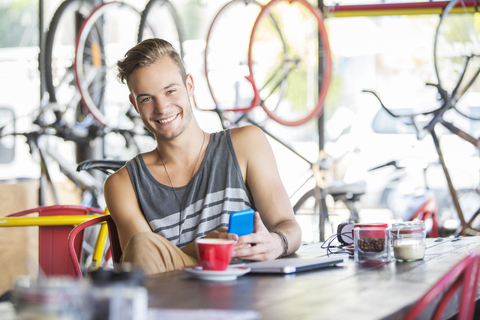 Porträt eines lächelnden Mannes mit Kaffee und Handy in einem Fahrradgeschäft, lizenzfreies Stockfoto