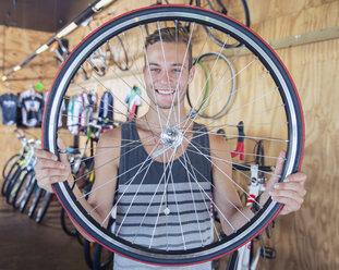 Porträt eines lächelnden jungen Mannes, der ein Fahrradrad in einem Fahrradgeschäft hält - CAIF13748