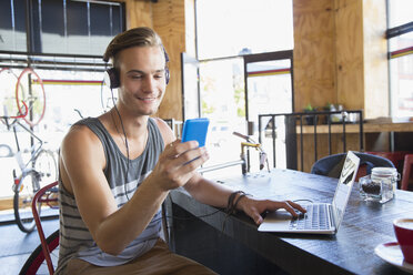 Lächelnder junger Mann mit Kopfhörern, der mit seinem Handy am Laptop in einem Café SMS schreibt - CAIF13749