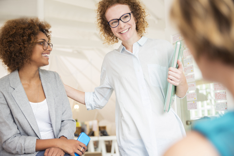 Frauen lächeln und sprechen im Büro, halten Smartphone und Laptop, lizenzfreies Stockfoto