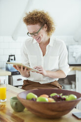 Porträt einer Frau, die in der Küche sitzt und auf ein digitales Tablet schaut, lächelnd - CAIF13808