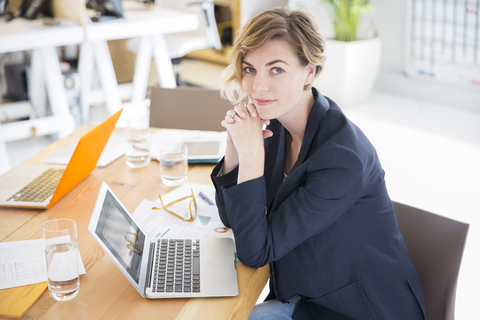 Porträt einer Frau am Schreibtisch sitzend mit Laptop im Büro, lizenzfreies Stockfoto