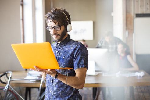 Casual businessman wearing headphones and using laptop in office - CAIF13840