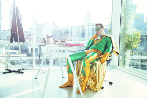 Gelangweilter Superheld, der am Schreibtisch im Büro sitzt, lizenzfreies Stockfoto