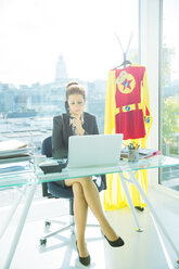 Geschäftsfrau bei der Arbeit am Schreibtisch mit Superhelden-Kostüm hinter ihr - CAIF13972