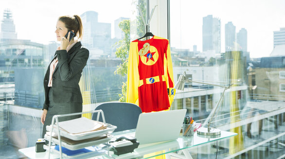 Geschäftsfrau im Gespräch am Handy mit Superheldenkostüm hinter ihr - CAIF13973