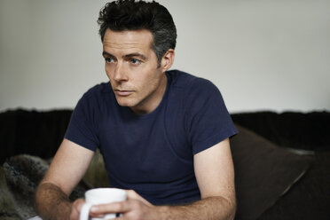 Man sitting on sofa holding mug looking sad - CAIF13980