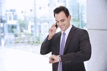 Lächelnder Geschäftsmann, der mit seinem Handy telefoniert und auf seine Armbanduhr schaut - CAIF14208