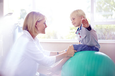 Physiotherapeutin hält Junge mit ausgestreckten Armen auf Fitnessball - CAIF14297