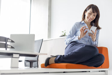 Lächelnde Geschäftsfrau, die mit ihrem Handy auf einem Kissen im Büro SMS schreibt - CAIF14759