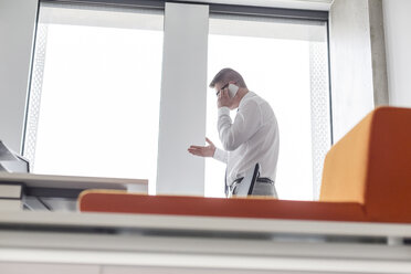 Geschäftsmann, der mit seinem Handy telefoniert und am Bürofenster gestikuliert - CAIF14762