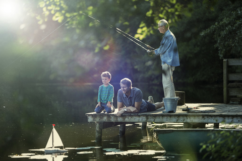 Junge angelt und spielt mit Spielzeug-Segelboot mit Vater und Großvater am See, lizenzfreies Stockfoto
