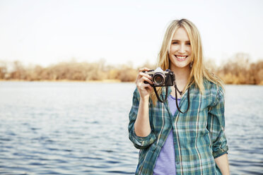 Portrait of woman holding camera while standing at lakeshore - CAVF06291