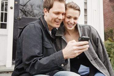 Couple looking at mobile phone while sitting outside house - CAVF06316