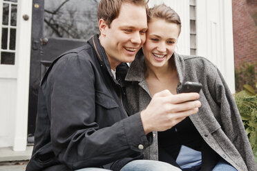 Paar schaut auf das Mobiltelefon, während es vor dem Haus sitzt - CAVF06316
