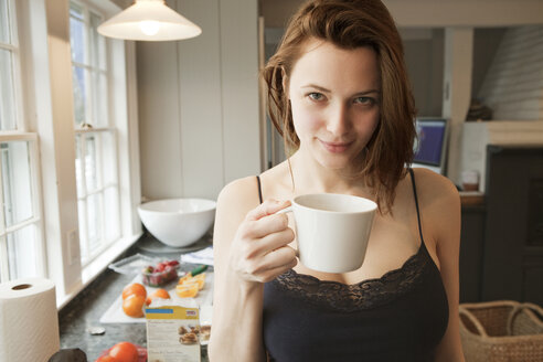 Portrait of woman holding coffee cup while standing in kitchen - CAVF06324