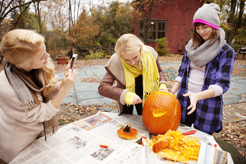 Frau beim Fotografieren, während Freunde am Tisch einen Halloween-Kürbis schnitzen, lizenzfreies Stockfoto