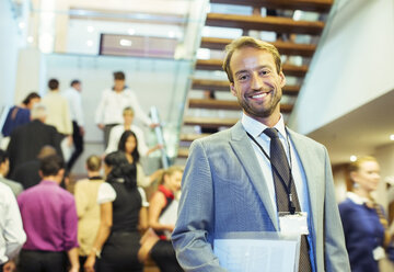 Porträt eines lächelnden Geschäftsmannes, der eine Akte hält und in der überfüllten Lobby eines Konferenzzentrums steht - CAIF15269