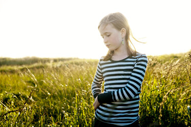 Girl looking down while standing in grassy field against clear sky - CAVF06523