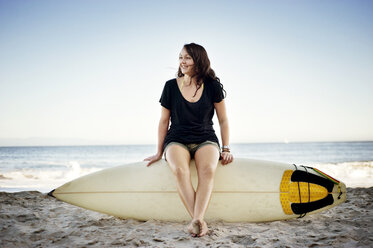 Frau schaut weg, während sie auf einem Surfbrett am Strand sitzt - CAVF06542