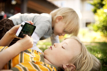 Junge benutzt Mobiltelefon, während sein Bruder im Garten spielt - CAVF06575